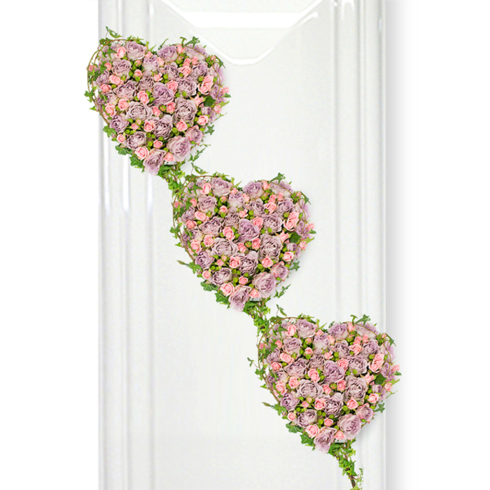 This image shows a beautifully crafted funeral tribute made up of three heart-shaped clusters of roses, artfully arranged to cascade in a diagonal line from the upper left to the lower right. Each heart is completely covered with miniature roses in soft, muted shades of lavender and dusty mauve, interspersed with pastel pink blooms that lighten the overall effect and add a touch of sweetness. The petals are tightly massed, giving the hearts a plush, cushion-like appearance, as if they might feel velvety and cool to the touch. Fresh green ivy is expertly wrapped around the outer contours of each heart and then trailed between them, its arching stems and glossy leaves forming graceful links that visually tie the trio together. This flowing greenery adds movement and a natural, almost garden-like feel, reminiscent of the climbing plants along Oxford's old stone walls and hidden courtyards. The entire design sits against a clean white background that includes faint vertical reflections, suggesting glass or a polished interior surface, which gives the arrangement a sense of light and space. The soft, even lighting reveals delicate shifts in colour across the roses, from dusky lilac to gentle blush, creating a peaceful, contemplative mood. This three-heart composition would be an especially meaningful tribute for a funeral or memorial service in Oxford, symbolising generations of love, the bond within a close-knit family, or the enduring connection between partners. It speaks quietly of affection, remembrance, and gratitude, translating those emotions into roses and trailing ivy.