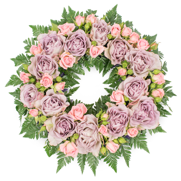 A serene funeral wreath is pictured from directly overhead, laid out on a clean white backdrop so that its flawless circular outline and careful symmetry are completely clear. The ring is densely filled with roses and greenery, creating the impression of abundance and thoughtful craftsmanship. Dominating the design are large, open roses in a refined palette of dusty lavender, antique mauve, and soft, greyish lilac, each bloom revealing tightly layered petals that spiral inward, like delicate paper folded again and again. These substantial roses are spaced in a gentle rhythm around the wreath, echoing the measured calm of an early morning walk along Oxford's tranquil side streets and college gardens. Between them, clusters of blush-pink spray roses weave through the design, their smaller heads offering contrast in both size and colour. Some of these little blooms remain as rounded buds, some are just beginning to unfurl, and a few are half-open, adding a sense of life in different stages and a tender, almost nostalgic charm. The flowers rest upon a thick bed of dark, glossy green fern fronds, likely leatherleaf or sword fern, whose finely cut, overlapping leaves form a continuous cushion behind the blossoms. This verdant framework creates a vivid border that defines the wreath's shape and brings out the softness of the purples and pinks. Subtle hints of deeper foliage and minute filler buds nestle in the shadows, lending quiet complexity. The even lighting highlights the textures of petal and leaf without glare, making the arrangement feel peaceful and contemplative. This is the kind of floral tribute families in Oxford might choose for a funeral, memorial service, or celebration of life, offering a gentle message of remembrance, gratitude, and enduring love.
