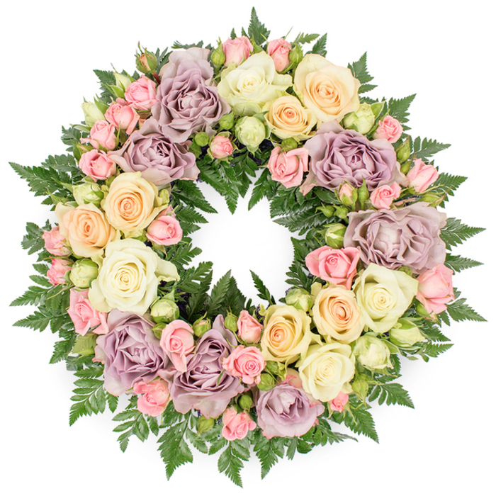 A classic circular funeral wreath rests visually against a pristine white background, its immaculate round shape immediately drawing the eye to the symbolism of eternity and continued remembrance. The wreath is lavishly composed, with roses packed so closely together that they form an unbroken carpet of petals, supported by a hidden framework of lush greenery. At the heart of the design are large, fully open roses in creamy white and soft ivory; their petals curl outward in layered spirals that catch the light and create a sense of quiet movement. Interlaced among them are clusters of petite blush pink spray roses, some still tightly furled, others just beginning to open, lending a sense of gentle transition and the passage of time. Soft peach and buttery yellow roses introduce a subtle glow that feels comforting rather than bright, while carefully placed dusty lavender and muted mauve roses add a unique, antique touch, as if echoing the stone tones of Oxford's old colleges. Below and around the blooms, fine, dark green fern fronds spread outward, their feathery texture providing a cool, verdant backdrop and peeking through like glimpses of shaded gardens off the Iffley Road or hidden courtyards in central Oxford. The lighting is calm and even, with no dramatic shadows, allowing each rose to be seen clearly and giving the arrangement a serene, contemplative presence. Crafted in a style familiar to local families arranging services from Botley to Headington, this wreath conveys deep sympathy, respect, and a wish for peace, making it a dignified tribute for a funeral, cremation, or memorial gathering. Prepared with the care you'd expect from an experienced Oxford florist, it offers a tender, visual expression of love and farewell.
