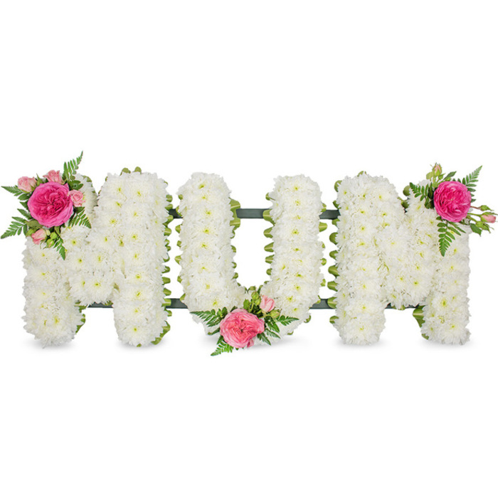 The image presents a tender funeral tribute spelling out the word MUM, positioned centrally against a bright, uncluttered white background that highlights every detail of the workmanship. The three letters are fashioned in tall block capitals, each one filled to the edges with countless white spray chrysanthemum heads. These petite flowers overlap in tightly massed layers, building a deep, plush texture that looks soft and pillowy, like a blanket of fresh snow. Their pure white tone suggests innocence, remembrance, and a gentle goodbye. Along the base of the letters runs a slim band of dark green ribbon, attached to a sturdy metal frame that connects the characters and makes it ready to rest on a stand for a funeral or memorial service anywhere in Oxford, from Cowley Road chapels to quiet cemeteries on the city's outskirts. To bring warmth and character, the florist has added vivid accent sprays: at the upper left corner of the first M sits a lush, ruffled fuchsia garden rose, surrounded by lighter pink spray roses and unopened buds, all threaded with delicate fern fronds that arc outward like soft green feathers. A similar grouping nestles into the lower centre of the U, cradling the middle of the word, and another mirrors the first, crowning the top right of the final M. These splashes of pink and green soften the formality of the white base, turning a simple word into a deeply personal gesture. It feels like a loving tribute a family might order from a trusted Oxford florist to celebrate the life of a devoted mother, whether for a church service, cremation, or quiet graveside farewell.