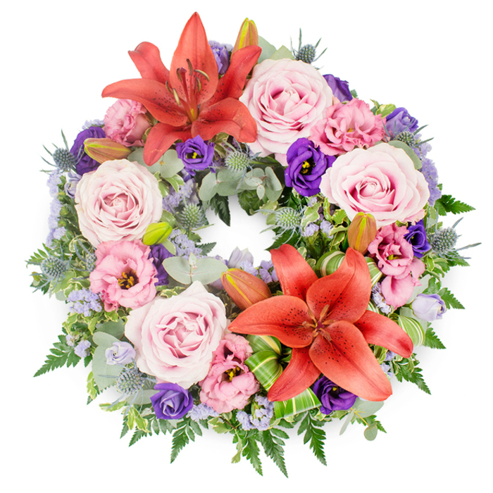 This photograph shows a meticulously crafted circular funeral wreath set against a pure white background, allowing every detail of the design to stand out clearly. The wreath feels both lush and carefully controlled, like a piece created for a thoughtful farewell at a memorial in central Oxford or a quiet gathering near the Botanic Garden. Two large coral-orange to reddish-orange Asiatic lilies serve as the main focal points, one displayed toward the upper arc of the circle and another positioned lower down. Their star-shaped blooms open wide, revealing intricate stamens at the centre, their petals gently curved and lightly textured, radiating a warm, dignified energy.
Between and around these lilies, an abundance of roses in soft blush and lavender-pink shades fills the ring, each flower fully open, with gently spiralling petals that suggest tenderness and grace. Intermixed are eustoma (lisianthus) flowers in two main tones: a delicate, almost pastel pink-purple and a rich, velvety deep purple. Their ruffled edges create subtle shadows and depth, echoing the layered history of the city's old stone colleges. Scattered throughout are tiny clusters of lavender-blue filler flowers, possibly statice, which add a light, airy haze of colour that softens the overall appearance.
Spiky blue-grey eryngium thistles punctuate the softer blooms, their bristled heads and slender stems contributing a cool, structural contrast. The floral elements rest on a deeply textured bed of foliage: silvery eucalyptus leaves, slender fern fronds, and dark glossy greens overlap to create a full, verdant frame. Several unopened lily buds peek from within the greenery, suggesting the gentle unfolding of future days. Near the lower lily, bright green leaves are folded and twisted into elegant shapes, highlighting the artisan skill behind the design. This wreath, ideal for same-day condolence delivery across Oxford, visually expresses remembrance, respect, and the unbroken circle of love.