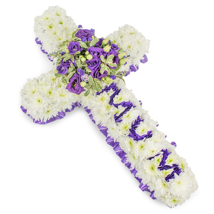 A large, meticulously assembled funeral cross presented against a clean white background: the body of the cross is entirely covered in tightly packed white chrysanthemum blooms, creating a soft, cushiony surface that conveys serenity and reverence. A contrasting border of deep, ruffled purple satin ribbon outlines the shape, its pleated texture framing the composition with formal grace. Along the vertical stem the name MUM is fashioned from smaller purple flower heads - possibly lisianthus or spray chrysanthemums - each letter clearly modelled so the tribute reads as a deliberate and loving message. The focal point at the intersection of the arms is a lush, opulent spray composed of dark violet and pale lavender roses, accompanied by pale purple lisianthus (eustoma) and variegated greenery with pale-edged leaves and darker centres, adding sculptural depth. Small, glossy green berries are woven through the central cluster, providing subtle contrast and tactile interest. The colourway - pristine white, soft lavender and deep violet - balances purity with dignity, creating an atmosphere of quiet reflection; a light, floral perfume seems implied by the dense petals. The arrangement's craftsmanship is evident in the even petal placement and harmonious colour transitions, qualities often requested for services at the University Church or private wakes near the Covered Market. Photographed in even studio light, this bespoke sympathy cross communicates respect and enduring affection with a restrained, elegant vocabulary of blooms and foliage.