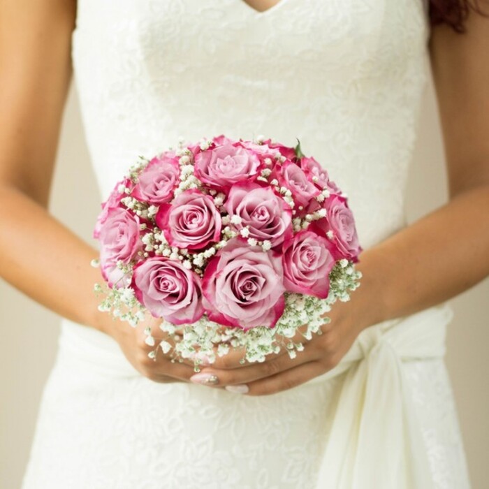 The photograph unfolds like a visual love letter to Oxford weddings-a close-up scene in which a bride, or perhaps a celebrant at one of the city's storied colleges, reverently holds a lush, rounded bouquet at her waist. The bouquet itself is a masterclass in balance and craftsmanship, formed by a tight cluster of medium-sized roses that blend seamlessly from creamy blush at the center to deep, jewel-bright magenta at the petal's edge, evoking the vibrant hues that might grace Oxford Botanic Garden in full bloom. Wispy white sprays of baby's breath nestle among the roses, their airy form providing cloud-like volume and setting off the vivid colours with a gentle, romantic counterpoint. Only the faintest hint of foliage peeks from the top, the stems expertly bound and hidden-much like the discreet elegance favoured by Oxford brides. The hands holding the bouquet, showcasing a subtle nude-pink shimmer on perfectly manicured nails, suggest careful attention to every detail. The sleeveless wedding gown is fashioned from fine lace, featuring a swirling floral motif and a graceful V-neckline-an ensemble that quietly references the refinement and heritage of Oxford's ceremonial traditions. A soft sash gathers on the right, infusing the overall composition with a sense of fluid movement. This image, elegantly composed and bathed in warm, natural light, speaks of meticulous preparation and classic romance-a scene fit for the city's radiant chapels or the lawns of Magdalen College, and a testament to the skilled florists at Flowers Oxford.