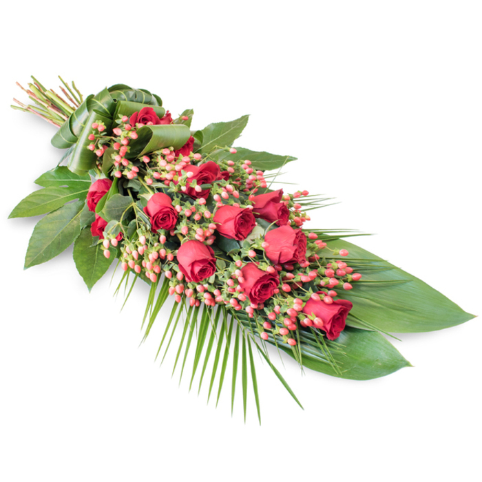 This image shows a long, elegant funeral spray arranged diagonally across a pristine white surface, the neutral background allowing every detail of the flowers and foliage to be seen clearly, as if laid out on a chapel floor before a service in Oxford. The spray begins at the upper left with tightly gathered stems, bound in a wide band of neatly folded green leaf that forms a smooth, natural cuff. From this binding point, the tribute extends gracefully downward and outward, forming an elongated teardrop or crescent shape. Along the spine of the piece sit approximately two dozen red roses, their petals a deep, velvety crimson. Some roses are still in bud, small and compact, while others are in mid-bloom, their petals gently spiralling open, and a few are fully open, displaying lush, layered centres. This mix of stages adds a sense of life and progression, as though capturing different moments in time. Between the roses, clusters of coral-pink Hypericum berries fill the gaps, the small, rounded berries providing a soft contrast in both shape and tone, almost like scattered beads of light among the blooms. Surrounding the central flowers, a generous base of dark green foliage supports the design. Broad, glossy leaves, possibly Ruscus or Salal, overlap to create a strong, dark framework, while finer, feathery palm fronds and slender stems radiate from the lower right, suggesting gentle movement and drawing the eye along the spray's length. The overall feel is of a classic, dignified sympathy arrangement, ideal for funerals, wakes, or memorials held in Oxford's historic churches and crematoriums, offering a heartfelt final tribute that balances sorrow, respect, and enduring love.