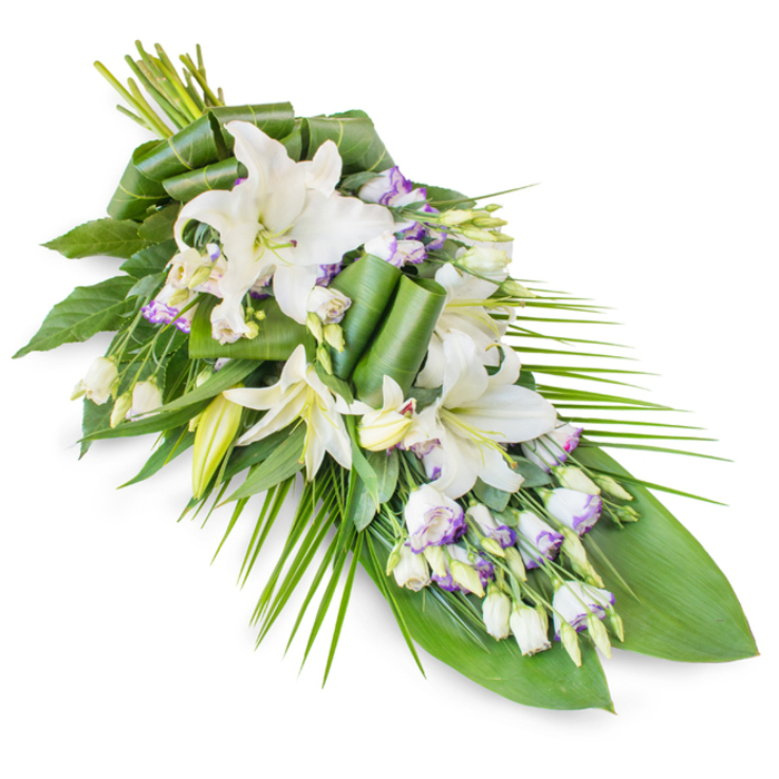A large, serene funeral spray designed in a classic elongated teardrop shape is shown against a crisp white studio backdrop, highlighting every nuance of petal and leaf. The arrangement is dominated by snowy white oriental lilies, placed along a diagonal that runs from a lush gathering of stems at the upper left to a gentle, tapering finish at the lower right-an elegant line often used for casket sprays and memorial tributes in Oxford churches. Some lilies are fully open, their broad petals unfolding like quiet wings around pale golden stamens, while others remain as smooth, elongated buds, subtly tinted with green at their tips. Nestled among these grand focal blooms are clusters of lisianthus flowers, each delicate cup of white edged with a soft whisper of lavender or light purple, adding just enough colour to soften the overall composition without disturbing its tranquil mood. Many lisianthus buds are still tightly closed, introducing fine texture and a sense of ongoing life. The flowers rest in a generous bed of assorted greenery: broad, dark leaves create a deep, velvety backdrop, while several are carefully folded, curled, or rolled into neat, contemporary shapes that betray a skilled florist's hand. Slender palm-like fronds extend outward, particularly from the right-hand side and lower portions, accentuating the spray's length and giving it a sense of gentle movement, like branches bending over the Cherwell on a quiet day. The lighting is soft and even, allowing the whites to appear luminous rather than stark. Altogether, the piece conveys peaceful remembrance and respectful farewell, an ideal sympathy arrangement for services held across Oxford, from Cowley Road to Summertown.