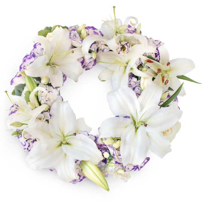 Set against a bright white background, this circular funeral wreath appears almost to float, its ring of white and lavender blooms forming a calm, comforting halo reminiscent of a quiet morning walk through Oxford's Botanic Garden. The wreath is luxuriously filled with white Oriental lilies, some fully open, some still unfolding their long, elegant petals. Their surfaces look silky and luminous, catching the light in soft highlights that draw the eye inward to the lily's green throat and prominent stamens dusted with amber-brown pollen. These commanding blooms are carefully spaced so their star-shaped faces overlap without crowding, creating a sense of balance and ease. Between and beneath them, lisianthus flowers add a romantic, layered texture; their gently frilled petals are white at the centre and brushed with lilac and purple along the edges, like watercolour strokes. Tighter buds and partially open lisianthus lend a sense of gentle progression, as though the wreath will continue to blossom through the course of the service. Discreet touches of deep and silvery green foliage peek through the petals, offering subtle contrast and helping define the wreath's generous, rounded profile. The colours are soothing rather than stark, ideal for a chapel service in Cowley, a crematorium ceremony, or a memorial gathering in a family home anywhere in Oxford. The unbroken circle quietly suggests eternity, while the combination of white and violet hints at purity, devotion, and spiritual reflection. Though the background is minimal and modern, the flowers themselves feel tender and traditional, creating a heartfelt tribute that honours the deceased with grace, beauty, and a sense of lasting peace.