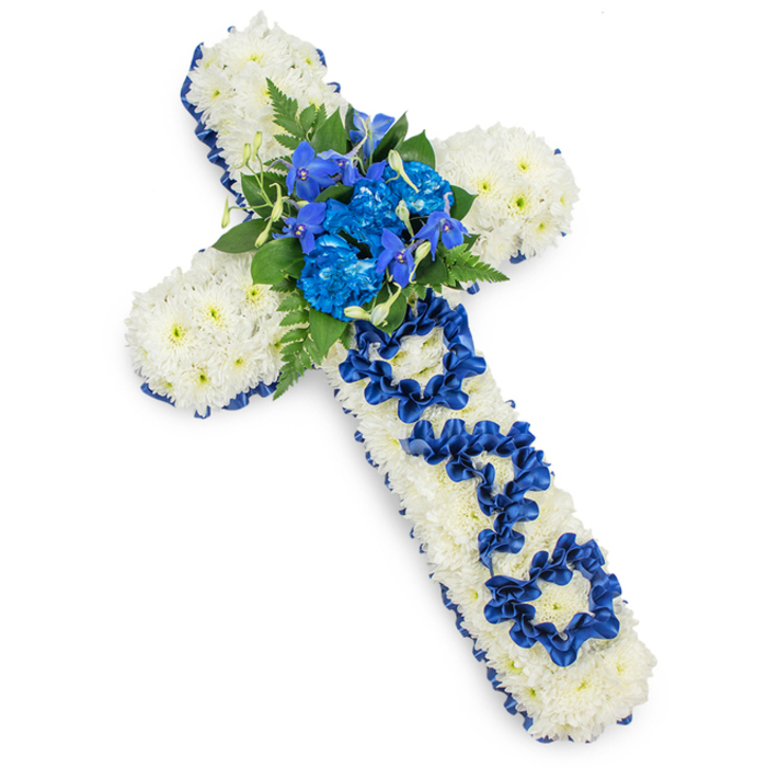 Set against a crisp white backdrop, a traditional Christian cross made entirely of flowers fills the frame, offering a quiet, reverent tribute. The body of the cross is composed of countless white spray chrysanthemum heads, placed so closely that no gaps of greenery show through, creating a thick, plush surface with a slightly domed contour. Each tiny bloom contributes to an overall impression of purity and calm, reminiscent of the soft light that falls over Oxford's college quads on a still morning. Around the entire outline of the cross, a rich royal blue satin ribbon has been carefully applied, its smooth sheen catching the light and providing a defined boundary between the white flowers and the surrounding space. This border curves gracefully along the arms and stem, lending the arrangement a polished, formal finish. At the central intersection, a vivid spray of blue flowers forms the main focal point: ruffled blue carnations or dyed chrysanthemums blend with spiky or upward-reaching blue delphinium or orchid florets, tucked into a nest of deep green leaves and feathery fern fronds. The greens break up the blues and whites, adding natural texture and a touch of woodland freshness. Running down the vertical arm, the word DAD appears in carefully looped royal blue ribbon, the letters gently resting above the chrysanthemum layer, clearly legible yet harmoniously integrated into the design. The composition is photographed from above with soft, shadowed edges, making it easy to imagine this cross laid respectfully beside a coffin or placed at a graveside service in one of Oxford's churchyards. It is a heartfelt, personalised funeral tribute that speaks of faith, gratitude, and cherished memories of a father.