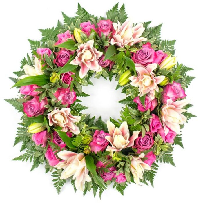 This photograph presents a meticulously crafted circular floral wreath, set against a bright white background that heightens every nuance of colour and texture. The wreath forms a perfect, unbroken ring, symbolising continuity and eternal remembrance, and is densely packed with fresh flowers that leave no empty spaces. At its core are luxurious roses in a rich pink palette: deep fuchsia and magenta blooms mingle with softer, mid-tone pinks, their petals layered in intricate spirals. Some roses stand proudly open, revealing velvety, ruffled centres, while others remain in tighter buds that lend variation and a sense of life in different stages. Interwoven with the roses are large double lilies, their petals broad, soft-looking, and gently curled at the edges. These lilies display delicate shades of creamy white, blush pink, and the faintest touch of pale peach, with a few petals showing slightly deeper veins or tinted edges. Several bright lime-green lily buds sit upright among the flowers, their smooth, tapered forms adding both colour contrast and a suggestion of blossoms still to unfold. Encasing the flowers is an abundant arrangement of greenery. Deep green fern fronds create a feathery, almost lace-like border, while other glossy leaves, some with subtle variegation, add body and depth. The interplay between the lush foliage and the vibrant blooms makes the wreath appear both full and balanced, with each flower seemingly cradled in green. The clean white background strips away any distraction, allowing the viewer to focus solely on the artistry and emotion embodied in the design. This type of wreath would feel at home at a funeral in Oxford, perhaps resting quietly by a graveside in Botley Cemetery or at a memorial gathering near Christchurch Meadow, offering a gracious, floral expression of sympathy and remembrance.