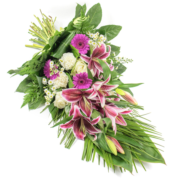This refined funeral sheaf is laid out against a pristine white background, emphasising its rich colours and thoughtful structure, much like a carefully composed scene in one of Oxford's historic colleges. The arrangement is long and slightly asymmetrical, flowing diagonally from the upper left, where the stems are neatly bound, down to a tapered point at the lower right. At the centre are several striking Stargazer lilies, their large, star-shaped petals painted in vivid fuchsia and deep magenta, each flared tip traced with white and scattered with burgundy speckles. A few partially closed buds, tinted green and pink, rise among the open blooms, adding height and a sense of living movement. Nestled alongside them are velvety magenta gerbera daisies, which bring bold blocks of colour, and clusters of creamy white roses that sit in various stages of opening, from tight bud to soft, cupped bloom. Ruffled white carnations fill the spaces between, adding gentle texture and a soothing, cloud-like softness. Fine sprays of tiny white daisy-like filler flowers thread through the composition, brightening the darker tones and creating delicate highlights. The flowers are cradled in an abundance of lush greenery: broad, shiny dark green leaves form the base and outline the shape, while feathery fern fronds and long, slender grass-like leaves extend outward, especially towards the bottom right, accentuating the cascading flow. This spray, crafted with the calm precision you'd expect from a seasoned florist serving Oxford's churches and crematoriums, feels both dignified and deeply personal. Ideal for a memorial gathering in Headington, a service in Cowley, or a quiet farewell at home, it conveys compassion, remembrance, and a graceful final tribute.