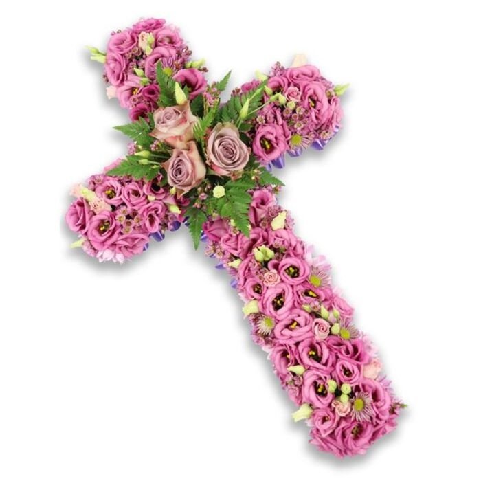 A lavish floral cross tribute, carefully arranged and photographed against a clean white background, is presented as a single, harmonious field of blossoms, every inch thoughtfully covered. The cross stands upright, its arms and base densely packed with lisianthus in soft pinks, gentle mauves, and muted lilacs, creating a tapestry of ruffled petals. The flowers are placed so closely together that they form a plush, almost cushion-like texture, with no stems or mechanics visible, only layers of delicate blooms. Some lisianthus show deeper rose-pink tones and slightly darker centres, adding quiet visual interest reminiscent of the varied pastel hues found on historic Oxford stone at sunset. At the centre where the vertical and horizontal arms intersect, three prominent roses form the focal point. These roses are larger than the surrounding flowers, choosing a dusty rose and light mauve colour that gently contrasts with the brighter lisianthus. Their petals unfurl in full, open spirals, suggesting tenderness and enduring love. Around them, feathery green fern fronds and smaller, polished leaves are tucked in to frame the roses, bringing a fresh, natural green accent. Throughout the cross, tiny clusters of pink and purple fillers mingle with palest cream and white blossoms, along with a scattering of light green buds, contributing to a layered, intricate texture. Along the edges of the cross, subtle glimpses of darker purple ribbon or fabric peek through, lightly defining its shape without competing with the flowers. The overall mood is serene and respectful, an ideal sympathy or funeral tribute for a service in an Oxford church near Summertown or by the riverside, capturing a sense of peace, remembrance, and gentle farewell.