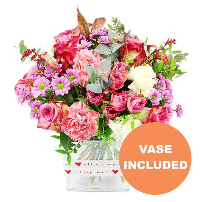 A richly coloured, carefully composed bouquet of mixed flowers sits in a clear, almost cube-shaped glass vase, set against a bright white background that gives the arrangement a clean, modern feel while allowing each bloom to shine. The eye is immediately drawn to the roses: large, velvety heads in deep fuchsia and magenta mingle with softer pink varieties, their petals layered like silk and some edged with slightly darker tones that add depth. Around them, generous carnations in shades of coral-pink, peach, and gentle blush unfurl their ruffled petals, creating a soft, cloud-like texture that feels both indulgent and welcoming, much like a warm afternoon spent in an Oxford college quad when the borders are in full bloom. Interspersed throughout are lively daisy-style chrysanthemums in lavender and light purple, with striking lime-green centres that pop brightly against the softer pinks and whites, joined by smaller creamy white blooms that lend brightness and balance. The bouquet is framed and filled by abundant foliage: rounded eucalyptus leaves in silvery-green, longer stems of darker greenery, and an occasional accent of reddish-brown leaves that adds a touch of seasonal richness, reminiscent of tree-lined paths through Christ Church Meadow. The arrangement is anchored in a transparent, rectangular vase wrapped at the base with a crisp white band. This band is decorated with a repeating motif of small, vivid red hearts and the handwritten phrase all my love, clearly suggesting romance, gratitude, or affection. In the lower right corner, an orange circular emblem carries the words VASE INCLUDED in bold white capitals, clarifying that the flowers arrive exactly as pictured, perfect for placing straight onto a dining table in Jericho, a student room near St Clements, or a reception desk on Beaumont Street. Crafted with care, it radiates warmth and celebration.