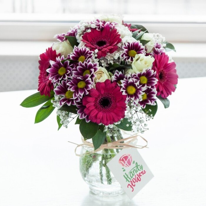 A striking mixed bouquet, clearly crafted by an experienced florist, sits centre stage in a clear, slightly ribbed glass jar-style vase, the kind you might find on a polished table in an Oxford kitchen filled with morning light. The flowers rise in a generous dome, with several large gerbera daisies in intense fuchsia and magenta commanding attention; their dark central disks provide a strong visual anchor amid the sea of colour. Interwoven among them are bicolour chrysanthemums, each petal tipped in rich purple that gradually fades into clean white, framing a small, golden-yellow centre that sparkles in the bright light. These daisy-like blooms add rhythm and texture, giving the arrangement a sense of playful depth. Creamy white spray roses are scattered thoughtfully throughout, some fully open in soft ruffles, others held in tight, promising buds, contributing a gentle, romantic tone. Fine sprays of baby's breath float between the larger flowers, tiny white blossoms creating a misty, almost ethereal layer that softens the bolder shades. Lush, deep green foliage supports the blooms, leaves peeking out at different angles and grounding the vivid colours. The vase gathers the stems neatly, and at its neck a piece of rustic twine is tied into a simple bow, from which hangs a tidy rectangular tag in light card, printed with a stylised pink rose emblem and elegant cursive lettering, though the specific wording cannot be read. The softly blurred white background gives the impression of daylight diffused through sheer curtains, typical of a quiet side street off Cowley Road or a calm study overlooking Christ Church Meadow. The bouquet feels fresh, joyful, and carefully curated, ideal for a celebration, thank-you gift, or everyday brightness delivered to homes and offices across Oxford.