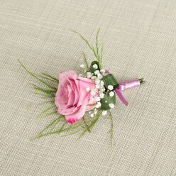 A delicately crafted pink rose boutonniere is displayed on a pale beige linen cloth, its subtle woven texture providing a calm, neutral stage that lets the flowers truly shine, much like a pressed shirt laid out before a ceremony near Oxford's historic Bodleian Library. At the heart of the design sits a single rose in full, fresh bloom, its petals layered in a gentle spiral. The innermost petals are a soft blush pink, gradually deepening to a richer, more vibrant pink toward the outer edge, where they curl slightly, giving the flower a sense of movement and softness. Encircling the base of the bloom are small sprays of white baby's breath, each tiny flower forming part of a fine, cloud-like ring that lightens the composition and adds a romantic, traditional touch often chosen for weddings and proms across Oxford. Behind the rose, slender blades of grass-like greenery splay outward in graceful arcs, their bright green colour adding contrast and an airy, contemporary feel. A broader, darker green leaf is folded with precision to wrap the lower portion of the stems, creating a neat, tailored base that suggests expert floral craftsmanship. The stems themselves are secured with a narrow satin ribbon in a harmonious pink tone, wrapped tightly and finished with a modest bow whose short tails rest naturally against the fabric. A small, silver-coloured pin sits discreetly at the base, indicating the boutonniere is prepared to be attached to a lapel-perhaps for a groom at an Oxford college, a guest arriving from Jericho, or a student dressing up for a prom night. Soft, diffuse lighting enhances the natural colours and gentle shadows, giving the entire piece a serene, intimate atmosphere.