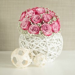 In this image, a carefully crafted round bouquet of pink roses and white baby's breath sits proudly in the centre, cradled by a spherical white woven vase on a spotless white surface. The roses form a generous dome, around fifteen to twenty blooms, each one opening fully so their layered petals can be seen, moving from a light, powdery pink in the centre to noticeably deeper pink, almost magenta, along the petal edges. Tucked between these roses, sprays of tiny white Gypsophila add softness and breathing space, like wisps of cloud drifting through a pastel sky, giving the whole arrangement a dreamy, romantic air. The vase is particularly striking: white strands, reminiscent of rattan or wicker, intersect in an open, basket-like structure, creating a delicate lattice that allows light to slip through and cast gentle shadows on the table beneath. The entire composition is placed on a smooth, bright white tabletop that subtly reflects the flowers and the base of the vase, suggesting a calm, tidy interior, perhaps a sunlit living room in an Oxford terrace near Jericho or Summertown. To the left of the bouquet, two decorative orbs made from intertwining pale wooden or fibre strips reinforce the natural, textural theme; one orb is noticeably larger, the other smaller, both slightly rustic yet modern in their simplicity. The background wall is a warm, creamy beige, softly textured and unobtrusive, which helps the pink roses and crisp whites stand out without distraction. The gentle, even lighting reveals every curve of the petals and every twist of the woven materials, evoking a feeling of quiet celebration, suitable for a Valentine's surprise, a birthday gift, or a welcoming arrangement on a hallway console in an Oxford home.