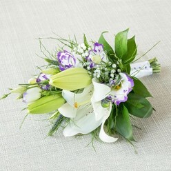 An elongated bridal-style bouquet of white lilies and purple lisianthus is arranged diagonally across a pale beige, gently textured fabric, the kind of subtle backdrop you might see on a polished table in an Oxford city-centre townhouse near Cornmarket Street. In the heart of the design, several large white Oriental lilies open fully, their curved petals glowing softly and revealing warm yellow-orange stamens dusted with pollen and tiny reddish freckles at the throat. Nearby, plump, light green lily buds stretch forward, not yet open but adding freshness and anticipation, as if the bouquet will continue to evolve after it leaves the florist's hands. Woven throughout, lisianthus blooms in creamy white with vivid violet and lavender edges provide a contemporary twist, their ruffled petals almost like silk, folding into subtle cups that add depth and movement. Fine sprays of white baby's breath scatter between the main flowers, giving a cloud-like, romantic haze that lightens the whole composition. The bouquet is supported by a generous framework of foliage: broad, glossy, dark green leaves that feel cool and substantial, softened by finer, feathery greenery and a few upright, spiky stems that suggest natural garden growth rather than rigid formality. At the tapered end, the stems are bound together with a smooth white ribbon, finished with tiny pearl-like beads that suggest a special occasion without overwhelming the natural beauty of the blooms. The lighting is soft and even, enhancing the creamy whites, sharp greens, and rich purples, creating a tranquil, elegant atmosphere. It feels perfect for carrying down the aisle in one of Oxford's historic churches, presenting as a thoughtful anniversary bouquet, or displaying as a refined centerpiece at a college dinner.