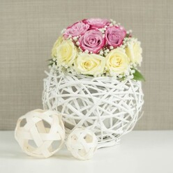A round, romantic centerpiece of roses and baby's breath rests on a white table, captured in soft, natural light that feels like it might be streaming through a townhouse window off Oxford's Iffley Road. The bouquet is carefully shaped into a dome, with roses closely clustered so that only their velvety heads are visible, no stems in sight. Blush-lavender and brighter fuchsia-pink roses mingle with creamy yellow and ivory blooms, the two palettes weaving together in a gentle contrast that suggests both sweetness and warmth. Threads of tiny white baby's breath drift between the roses, breaking up the colour blocks and giving the design a light, airy quality, as if the flowers are floating. This abundant crown of petals is cradled by a white spherical vase, made from fine twig-like branches or wicker painted a soft white and twisted into an open-weave globe. Through its gaps, you can glimpse the inner mechanics, hinting at the craftsmanship behind the arrangement. On the same crisp white surface, slightly to the left, sit two decorative rattan balls in natural, pale wood tones, their loose, looping structure echoing the main vase and adding a relaxed, organic contrast to the polished florals. The background is a warm, textured beige or light taupe, similar to a linen wallcovering in an Oxford café or boutique near the Covered Market, which gently frames the flowers without distraction. The mood is elegant yet inviting, perfect for a wedding table arrangement, a bridal shower in a North Oxford home, or a styled centerpiece showcasing timeless, handcrafted floral artistry.