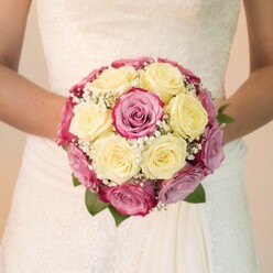 This close-up photograph captures the exquisite artistry of a bridal bouquet, the essence of Oxfordian romance reflected in every bloom. At the heart of the arrangement are lush, tightly clustered roses-some in creamy, buttery yellow, others in vibrant magenta with hints of soft mauve-reminiscent of the colourful displays at Botley Park in spring. Intertwined among the roses are ethereal sprays of white baby's breath, lending a light and airy texture that accentuates the fullness of the bouquet. Framing the bouquet, a few deep green leaves offer contrast and depth, while the bride's bare arms gently cradle this floral masterpiece. Her wedding dress, seen in subtle ivory-white tones, drapes softly and features intricately embroidered lace along the bodice, echoing the timeless elegance found in Oxford's historic college chapels. The lighting is gentle and evenly cast, illuminating each petal's velvety sheen and the delicate fabric's complexity. In the background, a muted softness keeps the focus entirely on the bouquet and the elegant lines of the bride's attire. The arrangement encapsulates a harmonious meeting of sophistication and local charm, suitable for any Oxford wedding, and stands as a testament to the skilled floristry available through Flowers Oxford-chosen with care, styled with love, and destined to become part of cherished wedding memories.