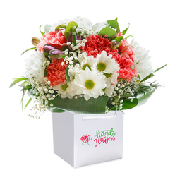 Bathed in soft, diffused light, a lush floral bouquet rises from a pristine white gift bag-inviting a sense of celebration and warmth that echoes through Oxford's winding lanes. At the bouquet's heart, richly textured carnations in shades of coral, peach, and scarlet unfold their ruffled petals, giving the entire composition a soft, velvety allure. Nestled among these are bright white daisies, each jewel-bright center bursting with life and echoing the freshness of springtime walks along the River Cherwell. Delicate sprays of snowy baby's breath interlace throughout, creating a dreamy, cloudlike effect that further enhances the bouquet's ethereal charm. A touch of purple from reserved, unopened buds on one side draws the eye and adds depth, the promise of more beauty to come much like Oxford's ever-changing gardens. Lustrous green leaves wrap the base, cradling each bloom with intention and providing a natural frame that highlights the careful artistry of the arrangement. The clean white bag is designed for effortless gifting-its ribbon handles and artisan branding suggest careful thought, much like a gift hand-delivered to an address in Summertown or delivered straight to an office in Oxford's historic heart. The presentation is expertly styled, perfect for landmark occasions or simply delivering a boost of joy on an ordinary day. Every detail reveals the expert touch of local florists who take pride in capturing Oxford's unique spirit through flowers.