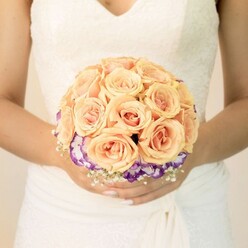This image offers a refined, mid-torso view of a bride holding her bouquet as if it were the heart of the day, captured against a softly lit, neutral background. Her wedding dress is a classic white or ivory shade, with intricate lace tracing the bodice and edging a modest V-neckline, while gentle folds of fabric at the waist and skirt fall in smooth, elegant lines. At the centre of the photograph, and of the moment, is a round hand-tied bridal bouquet, crafted with the kind of precision you'd expect from an experienced Oxford florist familiar with the city's historic chapels and riverside venues. The bouquet's core is a lush cluster of roses in shades of creamy peach, apricot, and blush pink, each blossom tightly cupped yet full, their petals unfurling in velvety spirals. Encircling this dome of roses is a striking ring of ruffled flowers, likely lisianthus, their petals saturated in deep purple that gently fades to white along the edges, adding drama and depth without overpowering the soft pastels above. Delicate white filler flowers, reminiscent of baby's breath, are tucked around the base, creating an airy border that lightens the overall composition. The bride's hands gently support the bouquet; her nails are neatly manicured in pale tones, with a subtle ombré or glitter accent on one thumb hinting at her personal style. The lighting is even and flattering, drawing the eye to the interplay of textures: the lace, the smooth fabric, the layered petals. It feels like a quiet pause just before walking into an Oxford ceremony, the bouquet radiating romantic calm and thoughtful craftsmanship.