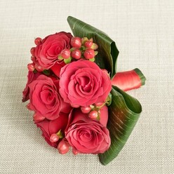 An elegant hand-tied bouquet of rich red roses and berries is displayed on a pale beige, softly textured fabric that could easily belong to an Oxford High Street boutique window. The arrangement is compact yet full, with five or six lush roses forming the dramatic heart of the design. Their velvety petals unfurl in tight spirals, each bloom at the perfect stage of opening, with deep crimson and scarlet tones offering a sense of passion and warmth. Tucked between the roses are small clusters of rounded Hypericum berries, glowing in shades of reddish-pink and cherry red, providing playful contrast and a hint of movement as your eye follows them around the bouquet. Supporting this central cluster, large, deep green leaves are artfully curved and folded, wrapping partially around the flowers like a protective cradle. Their smooth, shiny surfaces and prominent veins introduce a bold textural counterpoint to the softness of the petals. Around the stems, a narrow red satin ribbon is neatly wound, its subtle sheen suggesting expert, careful hands at work, such as those at Flowers Oxford when preparing wedding flowers for celebrations near the Bodleian Library or along St Giles'. Soft, even lighting gently illuminates every contour without glare, bringing out the richness of the colours and the luxurious feel of each element. The mood is undeniably romantic-perfect for a bridesmaid's bouquet at an Oxford college chapel, an anniversary dinner near the Covered Market, or a heartfelt thank-you gift sent to a home in Headington, where the arrangement's cool freshness and quiet elegance would make any room feel more special.