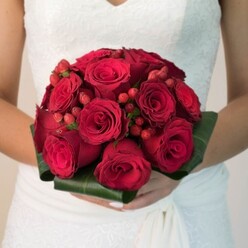 With the gentle warmth of Oxford's afternoon sun streaming through, this photograph draws its viewer into a world where tradition and romance walk hand in hand. The central focus is an exquisite bridal bouquet, artfully composed so that each velvety deep red rose appears curated for both visual drama and heartfelt meaning. Interlaced throughout are vivid, scarlet hypericum berries, their rounded forms offering playful texture and a subtle nod to the vibrant berries found at Oxford's Covered Market stalls. Lush, dark green leaves encircle the base, sculpted into a tailored collar, echoing the classic elegance of Oxford's Botanic Garden. Supported by the bride's neatly manicured hands, the bouquet radiates unity and abundance-its fullness suggestive of promises made and futures intertwined. She wears a white dress with delicate lace embroidery, its fabric softly cascading and catching the light filtering through nearby Radcliffe Square. The gentle scent imagined upon the air is sweet yet earthy, underscoring the passionate vows soon to be spoken. Flowers Oxford's craftsmanship ensures every detail feels deeply rooted in Oxfordshire tradition-perfect for a wedding day that seeks both timelessness and local distinction.