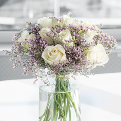 In this softly lit image, a graceful bouquet of creamy white roses and delicate lavender-pink filler flowers is displayed in a clear cylindrical vase, evoking the calm of a still morning on the banks of the Thames in Oxford. The roses are the heart of the composition-full-headed, open blooms with tightly layered, spiral centres that gradually unfold into gently ruffled outer petals. Their colour is not a stark white but a warm, soothing cream, tinged with ivory at the base, giving them a comforting, almost candlelit glow. Between these larger flowers, wisps of tiny lavender and blush-toned blossoms gather in airy clusters, adding texture and a dreamy, cloud-like effect, much like the drifting blossoms in University Parks in late spring. The filler flowers soften the strong, classic character of the roses and create a sense of hushed intimacy, suitable for moments of sympathy and quiet reflection. The bouquet is arranged in a tall clear glass vase filled with fresh water, where the cleanly cut green stems can be seen criss-crossing elegantly, grounding the composition in natural simplicity. The vase stands on a bright white surface that bounces the ambient light back onto the blooms, highlighting their freshness and subtle variations in tone. The background is softly blurred and minimalist-pale, luminous shades of grey and white-so that the flowers themselves become a calm focal point. It feels like an arrangement a local Oxford florist might place in a tranquil hallway in Headington or a peaceful sitting room near Cowley Road, bringing gentle solace to a grieving home.