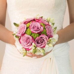 A meticulously arranged bridal bouquet takes centre stage, held closely by a bride within the storied backdrop of Oxford's city landscape, perhaps just steps from Magdalen College or a riverside garden. This lush arrangement bursts with the gentle allure of dusty rose and mauve-hued roses, their plush, velvety petals tinted at the edges with deeper fuchsia for added drama and depth. Bright lime-green pom-pom chrysanthemums nestle confidently amid the blooms, their spherical forms introducing a lively contrast reminiscent of Oxford's springtime parks. Draped around the outer rim of the bouquet, clusters of tiny, delicate white blossoms-likely fragrant sprays of rose or elegant freesia-soften the composition, imparting a timeless sense of bridal grace. Tiny green buds peek through the snowy fringe, infusing an organic vibrancy inspired by the city's lush surroundings. Illuminated by natural, diffused light, the intricate lace of the bride's gown glows faintly in the background-a gentle companion to the bold, gleaming floral colours. The bride's hands, manicured with a soft blush and a discreet sparkle, cradle the bouquet with reverence and joy. In this bouquet, crafted with an expert's touch-like that found at Flowers Oxford-the artistry reveals itself in every harmonious detail, making it a symbol of love, celebration, and the beauty that thrives in Oxford's wedding scene.