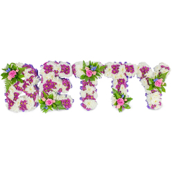 This memorial arrangement spells BETTY in large, sculpted capital letters, each letter formed from a thick, plush bed of white chrysanthemums that give the piece a soft, peaceful presence against the plain white backdrop. Nestled within those white heads, clusters of rich fuchsia-purple chrysanthemums map out the inner shapes and accents of every letter, their saturated tones creating a gentle but unmistakable contrast that brings the name into crisp relief. Around the verticals and curves a slender purple ribbon hugs the sides, lending a careful finish and a whisper of colour continuity. Small posies sit atop key points - on the B's upper curve, the top of the central T, and the Y's right arm - composed of blush pink roses in mixed stages of bloom, tiny sky-blue filler flowers such as delphinium or cornflower, and verdant fern fronds that add depth and a subtle forest-fresh fragrance. The roses' soft perfume and the faintly dusty scent of chrysanthemums combine to make the piece quietly fragrant without being overpowering. Crafted with funeral decorum in mind, the arrangement reads as both dignified and personal: a bespoke floral namepiece that carries tenderness and tribute. Locally, families in Oxford often choose arrangements like this for services at chapels off Botley Road or for display near the Covered Market entrance to remember a beloved family member, knowing the strong, simple colours and textures will convey sympathy and a lifetime of fondness.