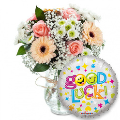 Soft morning light seems to settle over this comforting 'Good Luck' gift: a lush bouquet positioned slightly left of centre in a clear glass vase, its neck finished with rustic twine, the stems visible and neatly gathered below. As your florist I note the palette - pale peach and powder pink roses with tightly furled petals, paired with peachy-orange Gerbera daisies whose dark brown centres offer a warm focal point. Sprays of white Gypsophila tumble through the arrangement like airy lace, while compact clusters of white and lime-green chrysanthemum-like blooms add bright punctuation and crisp texture. Deep green foliage threads through the flowers, framing each bloom and lending a fresh, garden-cut scent that complements the roses' gentle perfume.To the right and overlapping the lower edge of the bouquet a round silver Mylar balloon gleams, its metallic face peppered with cheerful motifs: tiny hearts in blue and pink, stars in yellow and blue, and small multicoloured abstract shapes. Written across it in a playful, multicoloured typeface is Good Luck! - the letters bounce in green, orange, red and blue, and yellow smiley faces are playfully used as letterforms, lending friendly personality. The KALEIDOSCOPE brand mark is visible at the bottom rim of the balloon.This composition radiates encouragement and quiet celebration - perfect for sending to students before exams, colleagues starting new roles, or anyone facing a milestone. As Flowers Oxford often arranges for deliveries around Jericho, Summertown and the University Parks, the bouquet reads as both intimate and uplifting - a tactile, fragrant message of support made for neighbourhood moments.