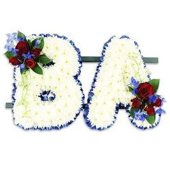 An intimate, expertly made standing tribute reads BA in bold uppercase form, each letter built from a dense carpet of small, immaculate white chrysanthemums that create a soft, layered surface you can almost feel beneath your fingertips. The perimeter of both letters is neatly outlined with rich royal blue satin ribbon, its gentle sheen lending a formal frame that accentuates the sculpted shapes against a stark white backdrop. Nestled on the upper left curve of the B is a lush cluster of deep red roses - some tightly closed, others opening into velvet cups - paired with sharp, cobalt-blue delphinium-like spikes that introduce a cool, lyrical contrast. A matching grouping on the lower right of the A repeats the pairing, tied together by darker green foliage and pale green trailing stems that drape with a delicate, organic grace. The two initials are mounted side-by-side on a low dark-green rectangular stand, creating a cohesive, freestanding memorial display that reads clearly from a distance. As a florist serving Oxford - from the Covered Market to Jericho and Headington - I recognise how colour and texture speak at a service: the white offers peace, the blue holds dignity, and the red whispers of deep affection. This arrangement, prepared with careful wiring and floral foam support, is suitable for funerals, memorials, and commemorative events where a personalised tribute is wanted; Flowers Oxford can prepare similar lettered tributes with local delivery across the city when requested.