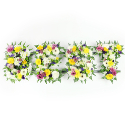 Presented against a clear, bright white backdrop, this horizontal tribute spells out "BEST" in four substantial floral letters, each one built from a tight mosaic of blooms and foliage that reads like a memory made manifest. The arrangement relies on a calming palette of crisp white chrysanthemums and cream gerberas, sunlit yellow roses and gerberas that seem to hold a warm afternoon glow, and small clusters of mauve to deep purple lisianthus which provide a gentle, reverent contrast. Vivid magenta-pink spray roses or carnations are woven through to offer small, joyful accents, while light-green and white filler flowers nestle into crevices to add softness and depth. Around each letter dark-green fern fronds fan out, creating a natural, almost lace-like border that frames the letters and tempers their solidity. Thin dark-green or metal support bars are discreetly visible where blooms cannot fully conceal the underlying structure, a sign of sturdy craftsmanship intended for display at a chapel, church hall, or funeral service. The head-on camera angle and evenly diffused lighting reveal petal textures, the faint leafy scent, and the fresh dampness of recently cut stems. As a florist's statement this piece balances bright, consoling colour with formal structure - an eloquent floral way to remember someone as "the best," suitable for friends and family seeking an uplifting but dignified tribute.