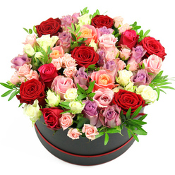 A striking floral hat box arrangement is shown in crisp detail against a clean white background, the simplicity of the setting drawing all attention to the abundant dome of roses. The flowers rise lushly from a sleek black round hat box edged with a slim red trim at the base, giving the design a modern, couture feel. Every inch of the container is brimming with roses in varied sizes and stages of bloom, from tight little spray clusters to large, fully opened heads unfurling layer upon layer of velvety petals. Deep crimson and rich red roses anchor the composition, their intensity softened by romantic blush pinks, playful fuchsia, warm coral and peach tones, and a scattering of lavender and pale purple roses. Creamy white and ivory blooms peek through, catching the light and creating a sense of movement across the dome. Delicate sprigs of fresh green foliage thread between the flowers, offering lightness and a gentle herbal freshness that you might associate with a walk along Oxford's Botanic Garden or through college quads after rain. The overall impression is one of generous luxury and polished craftsmanship, as though each stem has been placed with intention by an experienced florist who understands balance and colour harmony. This is the sort of arrangement that feels perfect for marking a proposal in central Oxford, a special anniversary dinner in Jericho, or a heartfelt thank-you to someone who has gone above and beyond. The bouquet's mix of passionate reds and soft pastels conveys romance, gratitude, and celebration all at once.