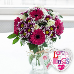 A bright, affectionate floral scene unfolds on a clean, light surface, as though set on a sunlit kitchen table in an Oxford townhouse. A rounded glass vase is filled with clear water, revealing neatly cut green stems that fan out into a generous dome of purple and white blooms. Bold magenta gerbera daisies steal the show, their wide petals and dark, velvety centres giving the bouquet a confident, modern feel. Nestled alongside them are eye-catching chrysanthemums in a rich, two-tone palette: deep plum-purple petals that gradually turn to white at the tips, encircling golden yellow hearts that glimmer like the late afternoon light over the River Thames by Folly Bridge. Soft, creamy white roses offer a gentle counterpoint to the intense colour, their layered petals curled inwards, suggesting quiet romance and tenderness. Sprays of fine white baby's breath thread delicately through the arrangement, adding texture and breathing space so each bloom can be admired. Lush green leaves frame the flowers, giving the bouquet a fresh, just-picked-from-the-garden character that feels at home anywhere from Summertown terraces to cosy homes near Cowley Road. Around the neck of the vase, a rustic loop of natural twine is tied, from which hangs a slim white gift tag decorated with a stylised pink rose and friendly green script that hints at thoughtfully chosen, hand-picked flowers. To the right of the vase, a shiny silver-rimmed heart-shaped balloon leans playfully into the composition. Its white centre is filled with colourful, patterned letters spelling out "Love n' Hugs 24-7" in joyful shades of pink, purple, and blue, dotted with small hearts. The softly blurred background, likely a sun-washed window, bathes everything in gentle light, creating an atmosphere of warmth and easy affection-ideal for cheering up a student in the city centre, celebrating an anniversary in North Oxford, or sending a spontaneous hug in flower form across town.