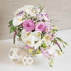 An elegant white wicker basket, generously filled with pink and white blooms, is captured in a bright, intimate close-up that feels like a table set for a celebration in an Oxford terrace near Cowley Road. The central stars are softly blushing pink roses, fully opened so their intricate spiral hearts are clearly visible, each petal curving outward in velvety layers. Between them drift large white Phalaenopsis orchids, their smooth, arching petals framing sunny yellow and soft amber centres, lending an air of contemporary luxury to the design. Around these statement flowers, frilled white Lisianthus with hints of lilac along the edges add delicate ruffles, while stems of pale pink stock spill gently over the side of the basket, suggesting a relaxed, romantic movement. Fine sprigs of white baby's breath lace through the arrangement, breaking up the larger heads with airy clusters that give the whole piece a light, cloud-like softness. Pockets of rich green foliage and emerging buds peek between the flowers, grounding the pastel palette with a fresh, natural contrast, as though the stems were just gathered from a cottage garden outside Oxford. The basket sits on a clean, white surface, where two small woven spheres in natural wood tones rest beside it, their interlaced strands mirroring the wicker and adding a subtle decorative echo. A neutral, fabric-textured backdrop in soft beige keeps the focus on the gentle play of colours and shapes. The composition conveys quiet joy and sophisticated romance, perfect as a wedding table centrepiece, an anniversary gift delivered across Oxford, or a graceful floral accent for a stylish home gathering.