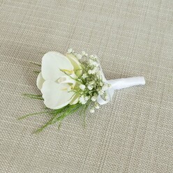 A refined white orchid boutonniere lies carefully placed on a neutral, light oat-coloured fabric, whose woven texture brings to mind rustic linen napery from an intimate Oxford reception. The central orchid bloom is a flawless Phalaenopsis, its wide, satin-smooth petals a luminous white that curves outward in gentle arcs. At its throat, a soft touch of pale yellow and cream adds a whisper of warmth, like the first light over the rooftops near Oxford's High Street. Around this focal flower, clusters of baby's breath gather in a soft, airy ring, each tiny white blossom contributing to a cloud-like halo that gives the piece a romantic, almost ethereal character. Fine, feathery sprigs of fresh green foliage thread through and beneath the gypsophila, their slender lines adding movement and a hint of woodland charm, as though picked from the edges of a path near Port Meadow. The stems of orchid, baby's breath, and greenery are drawn neatly together and bound with a pristine white satin ribbon, wrapped smoothly and finished with a discreet, tidy bow that holds everything firmly in place. The lighting in the scene is soft and diffuse, with no strong shadows, enhancing the calm, composed feel of the arrangement and letting the contrast between white petals, creamy ribbon, and lively green leaves quietly shine. This boutonniere feels designed for a groom or groomsman attending a classic Oxford wedding, perhaps in one of the city's historic colleges, where understated luxury, delicate scent, and careful craftsmanship combine to express love, commitment, and enduring elegance in a single, wearable floral detail.