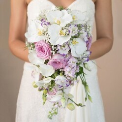 In this evocative scene, a bride stands against a softly muted, beige-toned backdrop, holding a lavish bridal bouquet that embodies the romantic heritage of Oxford. The focus is intimate, capturing only from her waist up as delicate light plays across the lacework of her strapless gown, hinting at a time-honoured tradition with a contemporary edge. Cascading downward in a graceful teardrop, the bouquet radiates a refined palette of whites, blush pinks, and tranquil lavenders-hues reminiscent of summer blooms at the University Parks or meadows near Christchurch. Centerpiece phalaenopsis orchids unfurl glossy white petals with sunbeam-yellow centers, their luxury contrasted by the soft, embracing fullness of pink garden roses, each petal plush to the touch. Amongst these, smaller lavender and purple stock blossoms add subtle notes of wild English fragrance, while crisp-edged purple lisianthus flowers and ethereal white baby's breath provide ruffled textures and a gentle cloud-like effect. Understated green leaves thread throughout, allowing the flowers and their natural beauty to shine-much like a quiet stroll along the Isis riverbank. The bouquet captures a sense of calm confidence, blending classical elements with airy movement. Each detail-from the shimmer of lace to the organic lines of trailing stems-whispers of romance and anticipation, perfect for weddings held in Oxford's historic venues. Crafted for the local bride who seeks sophistication and natural beauty, this bouquet is a true celebration of joy and artistry inspired by Oxford's elegant spirit.