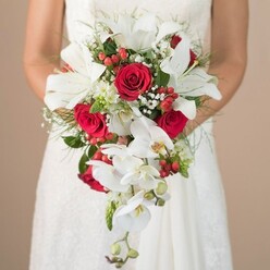 Captured in the soft, golden light of an Oxford afternoon, this wedding bouquet embodies the heartfelt joy and anticipation of a city-centre celebration. The composition focuses on the bride, whose figure is gently blurred in the background, drawing attention to the spectacular floral tapestry she supports just above her lap. At the heart of this shower-style bouquet, expansive white lilies open wide, their creamy petals reflecting natural light as their signature stamens stand proud. Dramatic touches come from the deep red, velvety roses whose lush petals nestle intimately among their white counterparts, offering a bold counterpoint of colour and emotion. The cascade unfurls with delicate white phalaenopsis orchids, their intricate lines and hints of yellow effortlessly draping downward as though mirroring the gentle flow of the Cherwell. Brilliant red berries-likely ripe hypericum-and airy baby's breath add joyful dashes of colour and ethereal texture, while fine sprays of soft greenery and deeper forest-shaded leaves frame the bouquet and evoke the lush green quads and leafy avenues of Oxford's neighbourhoods. The bride's white lace dress, visible at the torso and the curve of her arms, hints at timeless elegance and local craftsmanship, her posture conveying both tenderness and anticipation. Whether displayed at a stately Oxford wedding venue or exchanged along cobbled college lanes, this bouquet radiates tradition and sophistication-a perfect collaboration between inspired design and the historic charm of local floristry.