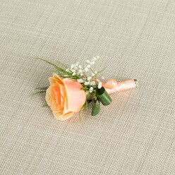A single peach rose boutonniere is displayed with quiet pride on a pale beige, linen-textured cloth, the subtle woven threads forming a soft, neutral backdrop reminiscent of a crisply pressed Oxford shirt. The rose itself is partially open, its petals moving from a warm, sunset peach at the centre to a paler, creamy tone at the edges, creating a gentle ombré effect that feels both modern and romantic. Nestled to one side, a cluster of dainty white gypsophila-baby's breath-adds an airy halo of tiny blooms, their star-like shapes dancing lightly around the rose. Slender green blades of foliage stretch out from behind, while two slightly larger, glossy dark green leaves curl protectively at the base, giving structure and balance. The stems are gathered tightly and wrapped in a smooth peach satin ribbon that mirrors the rose's colour, finished with a precise, understated knot that suggests practiced florist hands. Soft, even lighting glows across the arrangement, picking up the satiny sheen of the petals, the fine texture of the fabric, and the delicate shadows cast by each sprig of foliage. The boutonniere feels designed for a refined yet relaxed celebration in Oxford-perhaps a wedding in one of the colleges near Christ Church Meadow or a prom evening along the Cowley Road-where understated elegance is key. It conveys the quiet joy of a special day, ready to be pinned to a lapel and worn close to the heart, a small but meaningful floral accent thoughtfully arranged by a local floral studio.