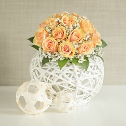 A refined floral display sits centered on a pristine white surface, drawing the eye with a domed bouquet of peach and apricot roses arranged in a modern, sculptural vase. Around twenty to thirty roses are closely nestled together, their petals tightly furled in places and gently opening in others, showing off a gradient of colour that moves from richer, sun-kissed orange in the middle of each bloom to soft, powdery peach and cream along the outer edges. Finely textured sprays of white baby's breath weave between the roses, creating a light, cloud-like halo that contrasts with the lush, velvety petals and adds a sense of airiness to the design. Here and there, small slips of deep green leaves peek through, adding a hint of woodland freshness and depth, much like the foliage you might see in the borders of Oxford's University Parks after a morning rain. The flowers are gathered in a spherical vessel made from slender white strands, intricately interlaced into an open lattice, so that the vase appears almost weightless, as though drawn with a single continuous line. In front of this main piece rest two smaller decorative spheres, crafted from the same pale material: one larger ball with a more structured pattern, and a second, smaller orb with a looser, more abstract weave, placed slightly off-center to give the composition a relaxed, contemporary balance. Behind the arrangement, a softly textured beige or greige wall keeps the background gentle and unobtrusive, allowing the warm floral tones and crisp white detailing to remain the focus. The scene is bathed in soft, even light that picks up every curve and shadow, suggesting a tranquil interior in an Oxford townhouse near Jericho, prepared for an intimate celebration, a romantic supper, or styled home decor photography. This thoughtfully crafted arrangement evokes quiet joy and understated celebration, showcasing the kind of careful attention to detail local florists bring to weddings, anniversaries, and same-day floral deliveries across the city.
