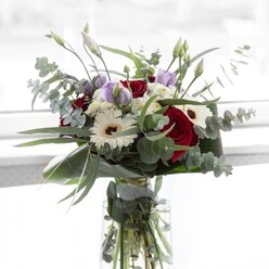 In the tranquil daylight that streams across Oxford's storied streets, a full and luxurious floral bouquet commands gentle attention. Lush, pillar-like red roses provide a sumptuous focal point, their petals lushly layered and inviting comparison to the vibrant displays found at Oxford's Botanic Garden. Flanking the roses, generous white gerbera daisies open out like miniature sunbursts, each centred with deep brown disks that hint at classic refinement. Softly coloured lisianthus in shades of pale lavender and gentle mauve are threaded among the larger blooms-some rising in slender, closed buds as if pausing before the full revelation of their beauty, suggesting hope and promise for special occasions such as engagements or graduations at the university. Tiny white filler flowers, airy as morning mist along the Thames Path, gently cluster to add dimension and texture. The ensemble is brought to life with the airy, silvery-green presence of eucalyptus, their round leaves releasing a subtle, crisp fragrance that mingles beautifully with the sweetness of rose. Lush, dark green leaves form the structure, cradling all within a pristine glass vase that exposes the fresh stems and pure water. This distinguished bouquet, available from local Oxford florists, feels equally suited to brightening a tranquil Jericho living room or offering a thoughtful gesture to friends celebrating in Headington, echoing the city's understated elegance and natural grace.