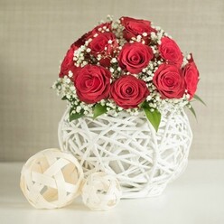 A lush, spherical cluster of red roses dominates the centre of the photograph, each flower in immaculate full bloom, their petals layered in tight spirals that create a luxurious, almost velvety canopy of colour. The roses, around a dozen or slightly more, are threaded with airy sprigs of white gypsophila, the baby's breath forming a soft halo of tiny blossoms that lighten the composition and heighten the sense of romance. Small touches of rich green foliage peek out between blooms, hinting at the natural stems hidden below and adding depth to the miniature landscape of petals. This radiant bouquet is cradled in a bulbous white vase with an intricate, open-weave structure, reminiscent of finely woven rattan or wickerwork. The lattice design wraps around the rounded form, letting glimpses of shadow show through the gaps and giving the impression of an artisan piece chosen with care. The vase rests on a pristine, pale surface that might be a modern dining table set for a reception in an Oxford college hall or an elegant home near Summertown. To the left, two cream-coloured decorative balls made from flat, interlaced strips echo the vase's texture, their differing sizes adding a gentle rhythm to the arrangement. Behind, a softly blurred, neutral wall in light beige or oatmeal tones keeps the focus firmly on the flowers. The overall mood is intimate and refined, like a wedding table arrangement ready to welcome guests, the quiet light suggesting an early afternoon celebration somewhere just off the High Street or by the dreaming spires.