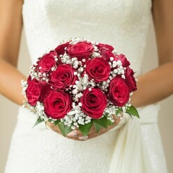 In this evocative moment on the edge of Oxford's dreaming spires, a bride's hands delicately cradle a domed bouquet of sumptuous, deep red roses, each rose unfolding its petals like verses in a love letter. The artistry is evident: every rose is nestled intimately alongside sprigs of dainty, snowy gypsophila, creating the impression of little constellations twinkling amid the crimson hues. Glossy green leaves provide grounding structure and a refreshing brightness, reminiscent of leaves glimpsed along the paths of Christ Church Meadow. The bouquet's passion meets pure grace as it rests against the bride's immaculate gown, which shimmers ever so slightly with the intricate patterns of lace or embroidery-its texture testament to the rich heritage of celebrations across Oxford. A soft ivory drape falls to one side, harmonizing effortlessly with the natural beauty of the florals. The scene is set in gentle, diffused light, the background fading to warm, neutral tones, evoking a hush of anticipation, like the first moments before saying "I do" in a centuries-old college chapel. This bouquet is more than floral artistry; it's the essence of Oxford romance-ideal for brides seeking a classic statement, or for articles on local wedding inspiration, floral design, or meaningful Oxford ceremonies.