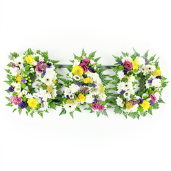 A moving floral arrangement spells out "DAD" in three bold, freestanding letters, the design captured against a spotless white background that highlights every nuance of colour and texture. Each letter is formed on a substantial base of rich green fern, the fronds overlapping in soft waves to outline the familiar shapes and suggest a natural, garden-like feel. Inside this leafy frame, the florist has densely layered traditional white blooms - generous chrysanthemums and wide-faced white gerbera daisies - so that the surface appears almost quilted, symbolising purity, respect, and quiet reflection. Nestled amongst these whites are smaller clusters of sunny yellow flowers, including yellow gerberas and dainty yellow spray roses, their cheerful tones adding a note of comfort, like a shaft of light through the solemnity of the day. To balance the palette, deep purple lisianthus and vivid pink or magenta spray roses are threaded thoughtfully through the tribute, creating pockets of rich colour that speak of enduring love and gratitude. Tiny lavender flowers and bright lime-green button chrysanthemums add extra texture and a subtle freshness. Slender dark green bars behind the letters keep them aligned in a neat horizontal row, but from the front they appear as a single, flowing word. This "DAD" tribute feels both traditional and deeply personal, the kind of bespoke funeral arrangement an experienced Oxford florist might prepare for a service in Jericho, a quiet farewell at Wolvercote Cemetery, or a memorial gathering for family and friends travelling in from across the city and nearby villages.