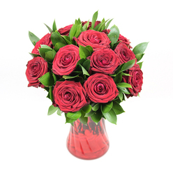 An opulent cluster of twelve to fifteen deep crimson roses stands proudly in a translucent red glass vase, photographed against a bright white background that lets the colours and textures sing. Each rose is captured at its most generous moment, the blooms fully open yet still full of life, with velvety petals curling out in perfect spirals that suggest both tenderness and strength. The roses' rich, ruby-red tones are intense without being harsh, softened by the studio lighting, as if echoing the romantic glow of an evening stroll along Oxford's High Street. Around and between the rose heads, dark green foliage-likely ruscus-fans out in glossy, pointed leaves, adding structure and rhythm to the design. The greenery's cool, polished surface contrasts beautifully with the softness of the petals, giving the arrangement a well-balanced, almost architectural silhouette. The vase itself is a deep red glass, its body gently curving from a sturdy base to a flared rim, and its subtle ribbed texture catches and refracts the light, creating quiet waves of shadow and highlight that complement the flowers above. Through the translucent glass you can glimpse the stems, carefully trimmed and arranged, reinforcing the sense of professional craftsmanship you would expect from a specialist florist serving the Oxford community. The pure white backdrop eliminates distractions, allowing every nuance of colour, from the darkest petal folds to the brightest green highlights, to stand out. Together, the roses, foliage, and vase form a timeless romantic gesture-ideal for anniversaries, declarations of love, or same-day surprise deliveries to homes and offices around Jericho, Summertown, or the city centre.