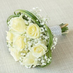 There's a serene, almost scholarly elegance to this hand-tied bridal bouquet, evocative of Oxford's enchanting blend of tradition and contemporary style. Soft, velvety roses in creams and buttery whites unfurl their petals, bathed in cool natural light like the delicate blooms in a Botanic Garden greenhouse. These roses are thoughtfully clustered with puffs of white gypsophila, their lacy texture dancing around the roses in a pattern as intricate as the ironwork on High Street's historic balconies. Sweeping arcs of slender, deep-green foliage provide both structure and whimsy, reminiscent of the graceful curves found along the banks of the Cherwell. The bouquet's stems are secured with a luminous silver-grey ribbon, studded with neat rows of tiny pearls-a tactile finish that echoes the refined detail Oxford is known for. There's a sense of gentle warmth and anticipation, as if this bouquet is awaiting its turn to be carried through the streets of Jericho or celebrated at a reception in the heart of Summertown. Crafted with artistry and intention by Flowers Oxford, this arrangement speaks to lasting romance and quiet sophistication and is available for delivery across every cherished corner of the city.