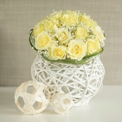 A luxurious floral centerpiece glows with quiet elegance, built from dozens of pale yellow roses cupped together in a flawless dome, their petals layered like soft satin. Nestled delicately between the roses, airy tufts of white gypsophila add a misty, romantic texture, softening the overall shape and evoking a bridal veil. At the base, a sleek ring of deep green foliage encircles the bouquet, defining the lower edge and adding a fresh contrast that highlights the luminous pastel tones. This artful arrangement is cradled in a sculptural white woven vase, its basket-like lattice of slender strands creating a sense of lightness and movement, as though the flowers are floating. The vase stands on a polished white surface, where two decorative spheres made from pale rattan-style strips are placed thoughtfully alongside, their open, geometric weave reflecting the pattern of the vase. Gentle, diffused light washes across the scene, picking up the creamy yellows and cool whites, while a soft beige background, similar to the mellow stone you see around Oxford's colleges, keeps the focus fully on the flowers. The arrangement feels serene and impeccably composed, ideal as a wedding table centerpiece at a chic Oxford venue near the Bodleian or along St Giles, or as a refined decoration for a civil ceremony reception in Headington. The image conveys the quiet confidence of expert design and fresh, carefully selected blooms, arranged to bring an atmosphere of joy, grace, and thoughtful celebration to any special gathering.
