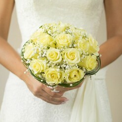 In this tender Oxford bridal moment, a lush bouquet of soft yellow roses is showcased with heartfelt artistry, lovingly held by a bride whose carefully manicured hands and shimmer of a wedding band evoke love's quiet promises. Each rose, tightly layered within the domed bouquet, reveals petals curling outward with gentle confidence, their golden hue reminiscent of early spring mornings along the River Cherwell. Wisps of baby's breath interweave amongst the blooms, adding a touch of airy delicacy that brings depth and movement, like the mist rolling across Port Meadow at dawn. At the base, looping bands of slender greenery encircle the stems, their smooth, dark texture mirroring the well-tended hedgerows that line Oxford's winding lanes. The bride's dress glimmers in soft focus-a vision in white, with hints of textured fabric exploring the play of sunlight on embroidered details. The background is artfully blurred, ensuring each floral detail remains the center of attention. This bouquet, expertly curated for Oxford wedding celebrations, bursts with both warmth and subtle fragrance, inviting memories of intimate vows and shared hopes-a true symbol of elegance for occasions where tradition and new beginnings entwine.