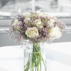 A full, rounded bouquet of white roses and lavender statice is captured in soft natural light, as if placed near a sunlit window in an Oxford terrace close to Jericho. At the heart of the arrangement, around a dozen creamy white roses open with quiet confidence, their petals layered like folded silk, some blooms more tightly furled while others gently unfurl to reveal a warm, creamy centre. Weaving in and around these classic flowers are airy clusters of tiny, pastel lavender statice, their cloud of miniature blossoms lending the design a lighter, almost misty texture. The statice forms a soft halo around the larger rose heads, giving the bouquet a balanced, almost floating appearance. The flowers are gathered in a simple, tall cylindrical glass vase, the transparent sides revealing straight green stems fanning slightly as they meet the base, all immersed in clear water that glints where the light touches it. The vase rests on a pristine white surface that reflects the arrangement's colours and intensifies the overall feeling of cleanliness and calm. The background fades into a gentle blur, suggesting an indoor space filled with daylight but free of distractions. The mood is tranquil and refined, well suited to occasions where a gentle presence is needed-a condolence bouquet for a quiet home near Cowley Road, a graceful anniversary gift, or a sophisticated accent on a reception desk. Crafted with the care you'd expect from an established Oxford florist, the arrangement conveys soft romance and thoughtful simplicity without ever feeling overstated.