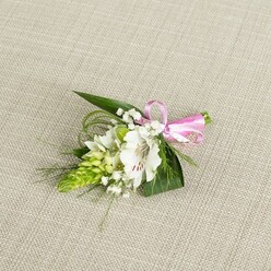 A refined floral boutonniere rests on a pale beige, linen-like cloth that recalls the soft, neutral tones of Oxford's honey-coloured buildings, giving the small arrangement a serene, gallery-style setting. At its heart, a single white alstroemeria, or Peruvian lily, opens gracefully, its petals a clean ivory that shifts to a creamy warmth towards the centre, where fine reddish-brown streaks radiate delicately from the throat. The stamens, tipped with light pollen, add a subtle point of focus. Just to its left rises a compact, cone-shaped spike of vivid green buds, tightly stacked and slightly glossy, introducing a striking contrast of texture and springtime freshness. Around and between these focal elements, airy clusters of white baby's breath appear like tiny stars or drifting clouds, softening the lines of the design and lending an almost weightless feel. The structure is anchored by several dark green, pointed leaves, firm and smooth to the eye, which cradle the blooms, while finer, thread-like blades of bright green foliage arc outward, suggesting gentle movement, much like a breeze over Christ Church Meadow. On the right, the stems are neatly finished with a bright pink satin ribbon, tied into a small, confident bow that catches the light with a subtle sheen and injects a joyful accent of colour. The piece is evenly lit, with no harsh shadows, so that every nuance of petal texture and foliage is clear. The overall mood is one of fresh, polished celebration, ideally suited to a groom or prom guest in Oxford seeking a simple yet elegant floral detail that speaks of care, craftsmanship, and quiet romance.
