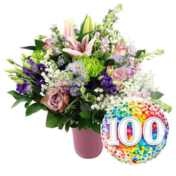 Set against a crisp, uncluttered white backdrop, this image showcases a sumptuous anniversary bouquet and a gleaming milestone balloon, carefully styled as if prepared for delivery to a cherished Oxford home near Jericho or Headington. The flowers spill generously from a ribbed, matte pink vase, the soft blush tone of the container echoing the romantic hues within. At the centre and rising above, graceful pink Oriental lilies catch the light, some fully open to reveal their star-shaped petals and prominent stamens, others still in elongated buds that hint at days of beauty yet to come. Around them cluster soft pink and deeper mauve roses, their layered petals suggesting both elegance and nostalgia. Between these classic flowers, rich purple lisianthus and sprays of lavender asters with tiny yellow eyes weave a ribbon of cool, soothing colour, while vivid lime green button chrysanthemums punctuate the arrangement with a contemporary twist. Wisps of white baby's breath float throughout like tiny clouds, softening edges and adding a sense of airiness. Deep, glossy green leaves and stems anchor the bouquet, giving it volume and natural depth as though freshly gathered from a well-tended garden off the Cowley Road. To the lower right of the arrangement, a round foil balloon catches the eye with its mirrored sheen and lively confetti pattern in bright rainbow shades. Dominating its centre, clear white "100" numerals with a subtle shadow effect proclaim a momentous centennial. Together, bouquet and balloon create an uplifting tribute, perfect for marking a 100th birthday, long marriage, or historic Oxford anniversary.
